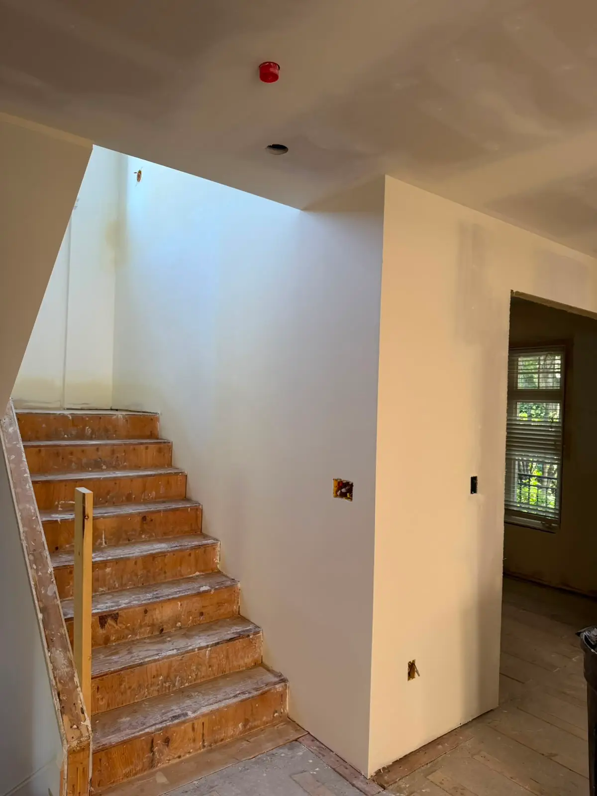 Stairwell with plywood treads and finished drywall walls, opening through to a room beyond.
