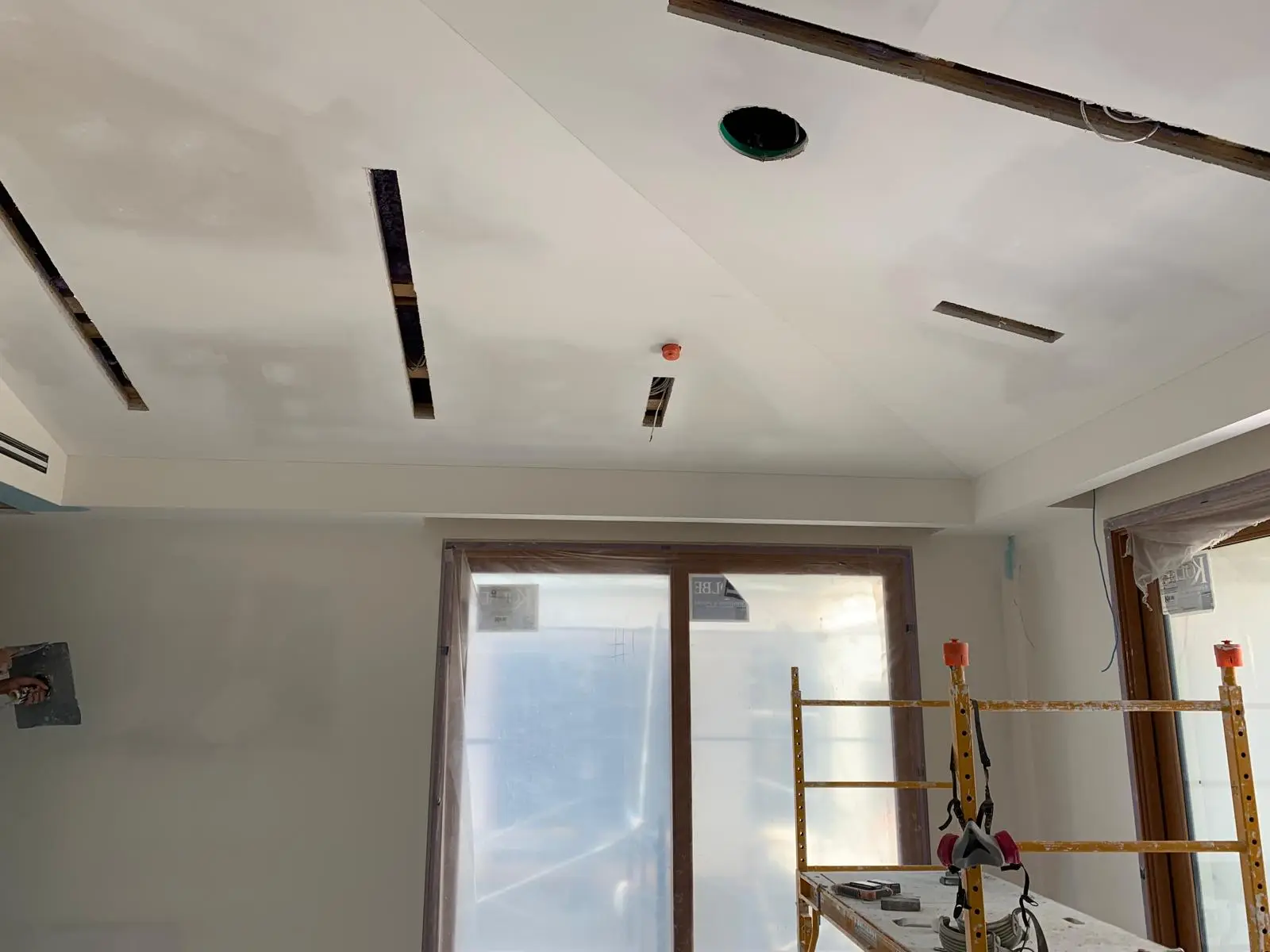 Tray ceiling with linear slot openings and pot-light cutouts above a sliding door wall, scaffold in the room.